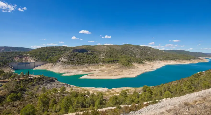 El pantano de Sacedón, con su agua turquesa, se extiende entre colinas verdes bajo un cielo azul.