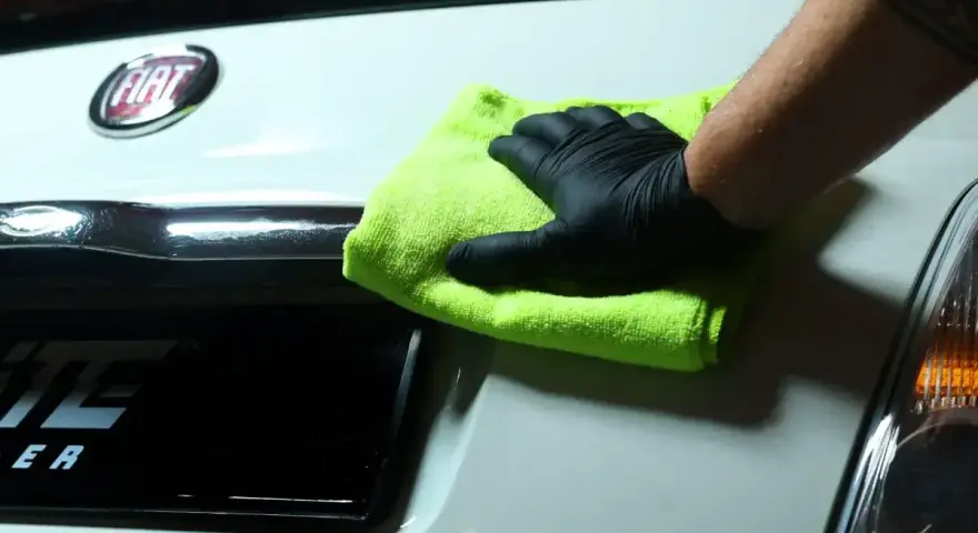 Ręka w czarnej rękawiczce z zieloną mikrofibrą czyści biały lakier Fiata. Idealne rozwiązanie, czym odtłuścić lakier przed polerowaniem.