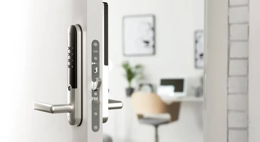 Modernt digitalt låssystem på en vit dörr. I bakgrunden syns ett kontor med skrivbord och stol.