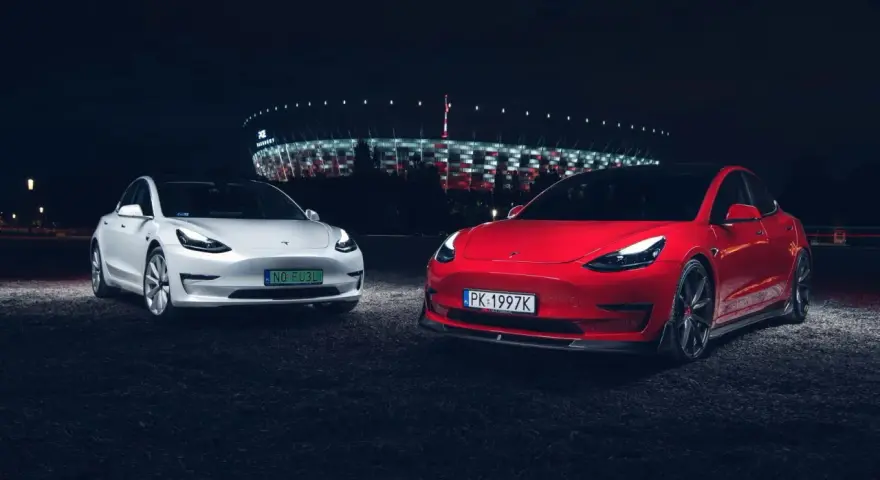 Dwa modele Tesla, biały i czerwony, zaparkowane nocą przed oświetlonym stadionem.