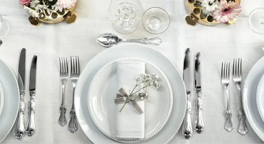 Ett elegant dukat bord med vita tallrikar, servetter dekorerade med blommor och vackra, silverfärgade engångsbestick.