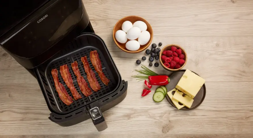 Knuspriger Bacon im Airfryer, daneben Eier, Beeren, Käse und Gemüse für ein perfektes Frühstück.
