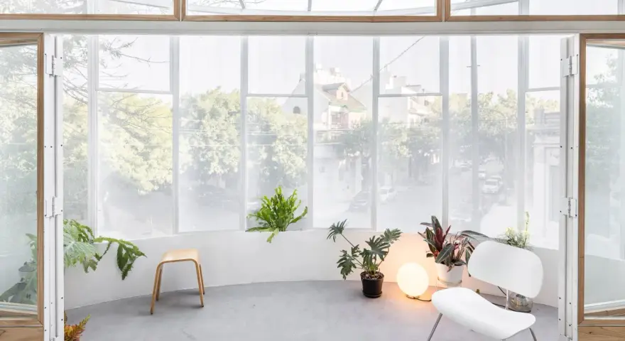 Interno luminoso con piante, sedia bianca e sgabello in legno, affacciato su una strada alberata da una veranda balcone.