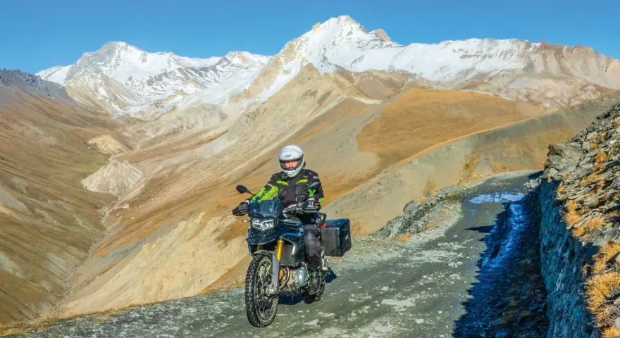 Un motociclista avventuroso affronta un sentiero impervio con la sua jafferau moto, circondato da maestose montagne innevate e paesaggi aridi.