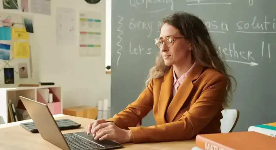 Nauczyciel początkujący w okularach pracuje przy laptopie w klasie. Na tablicy napisy po angielsku.