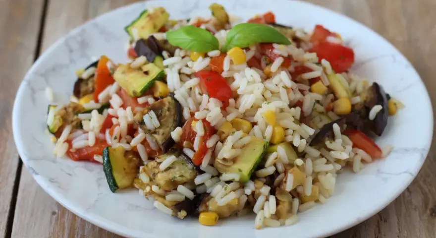 Un piatto colorato di insalata di riso vegetariana con zucchine, melanzane, pomodori e mais, guarnito con basilico fresco.