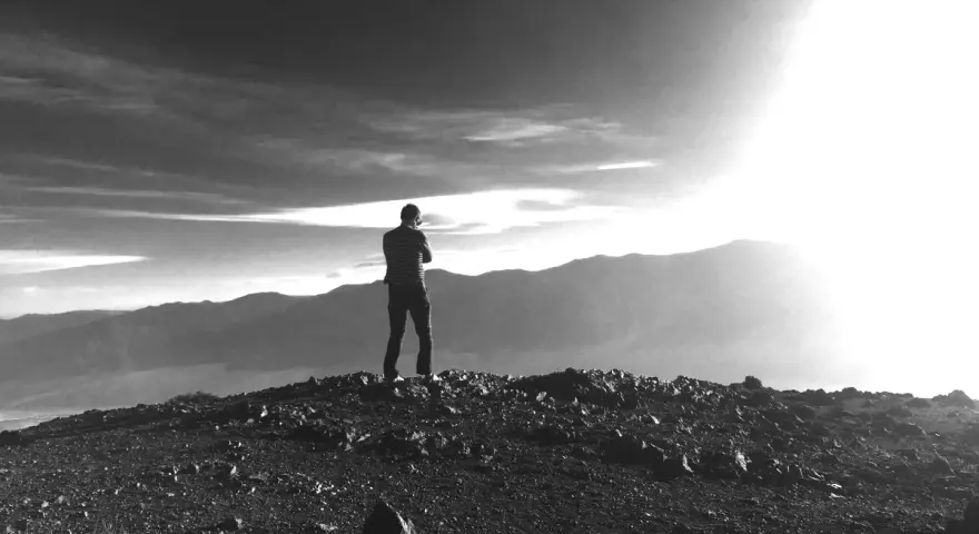Ein Mann steht auf einem felsigen Gipfel und blickt in die Ferne. Die Sonne bricht durch die Wolken und beleuchtet die Berge. Vielleicht fragt er sich, was ist Weisheit.