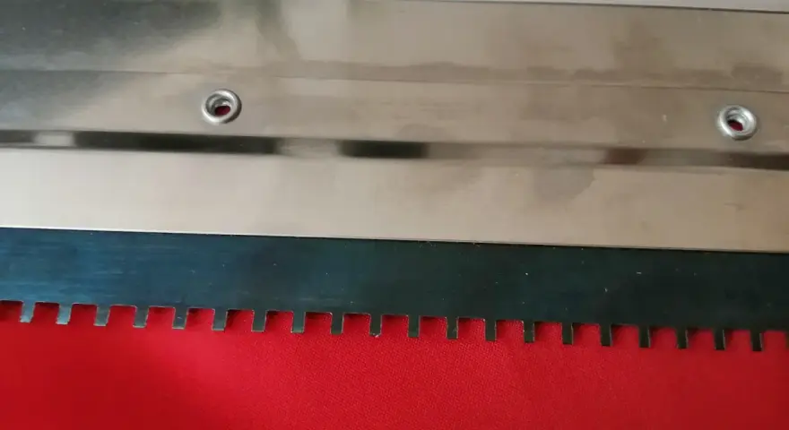 Metalowa rakla z ząbkowanym ostrzem, gotowa do pracy na czerwonym materiale.