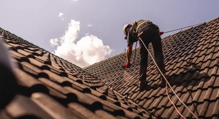 Pracownik w kasku i uprzęży zabezpieczającej wykonuje renowację dachu, natryskując dachówkę.