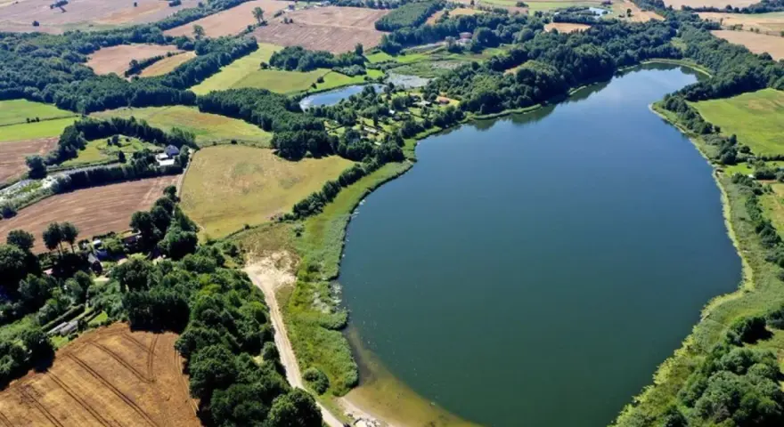 Malownicze jezioro Marszewo otoczone zielenią i polami. Widać piaszczystą plażę z pomostem i kilka samochodów.