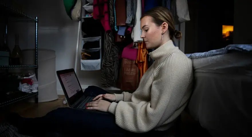 Ung kvinna sitter på golvet och jobbar på sin laptop, hon kombinerar studera på distans och jobba samtidigt.