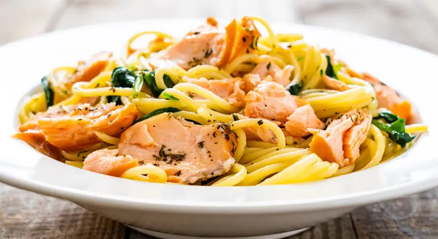 Deliciosa pasta con salmón desmenuzado y espinacas, servida en un plato blanco sobre una mesa de madera.