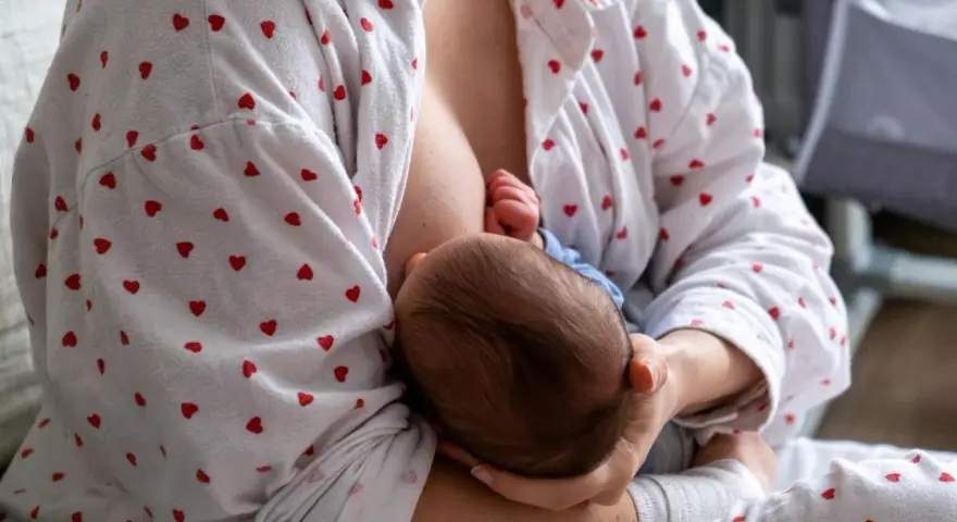 Matka karmi piersią dziecko. W tle widać łóżeczko. Zatkany kanał mleczny to częsty problem.