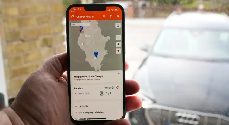 En hand håller upp en mobil med appen ChargeFinder som visar en karta över laddstationer för elbil. En laddapp visar en laddare nära en elbil.