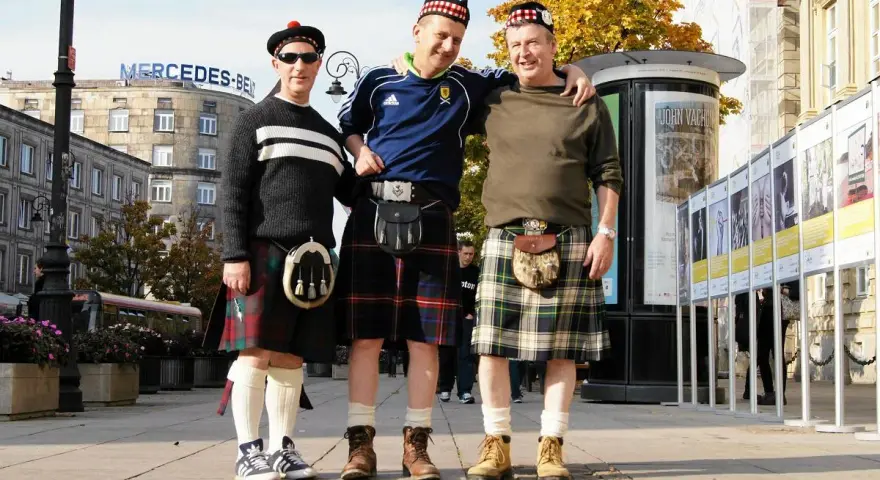 Trzech mężczyzn w tradycyjnych szkockich spódnicach, czyli kiltach, pozuje do zdjęcia na tle miasta.