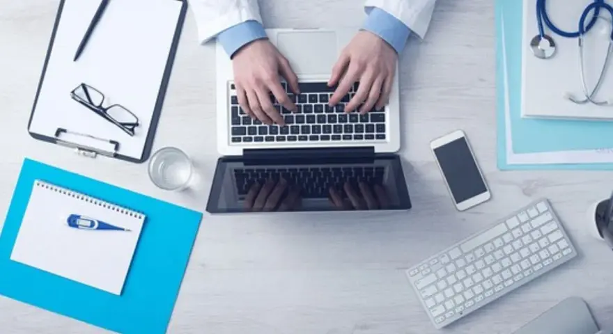 Lekarz pisze na laptopie, obok stetoskop, recepta, telefon i termometr.