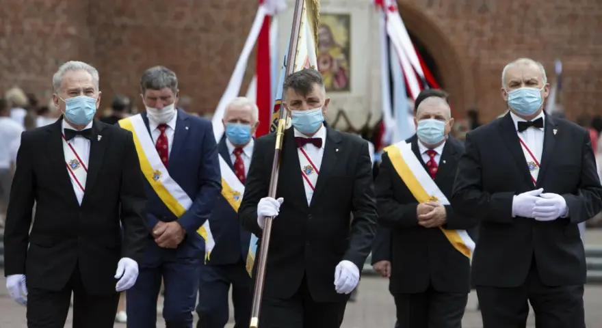 Rycerze Kolumba w garniturach i maskach maszerują z flagą.