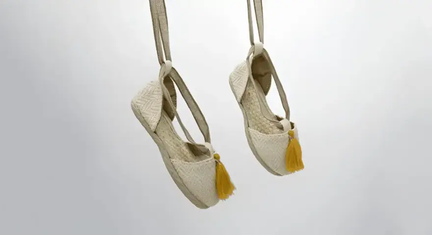 Dos alpargatas de tela beige con pompones amarillos cuelgan de tiras, mostrando su diseño veraniego.