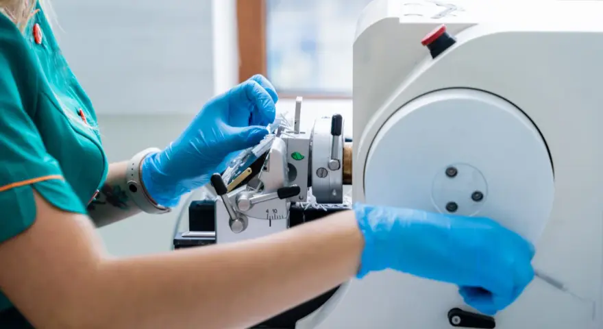 Ręce w niebieskich rękawiczkach pracują przy mikrotomie, przygotowując próbkę tkanki do badania. Może to być kluczowe dla diagnozy, czy wykryty guz jest złośliwy.