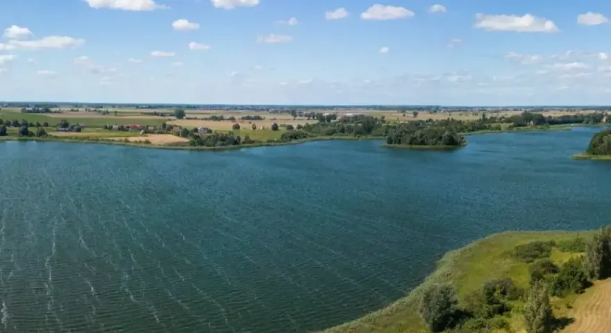 Malownicze jezioro Lednica z wyspą pośrodku, otoczone zielenią i polami. Błękitne niebo z białymi chmurami.