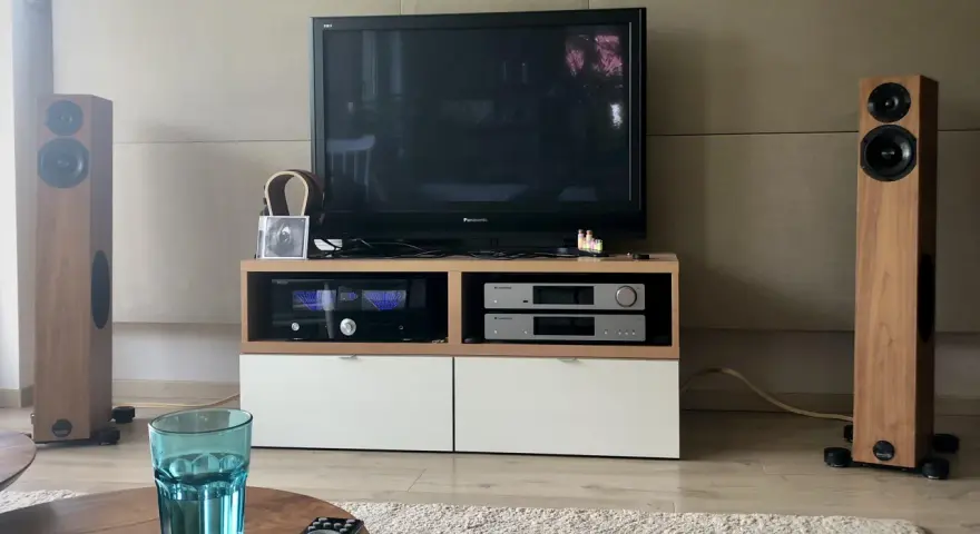 Dwie kolumny Audio Physic Virgo w drewnianej okleinie, telewizor, sprzęt audio i szklanka wody na stoliku.