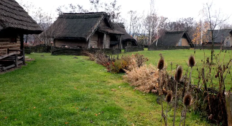 Park archeologiczny Osada VI Oraczy. Drewniane chaty z krytymi strzechą dachami, otoczone plecionymi płotkami, na tle jesiennej zieleni.