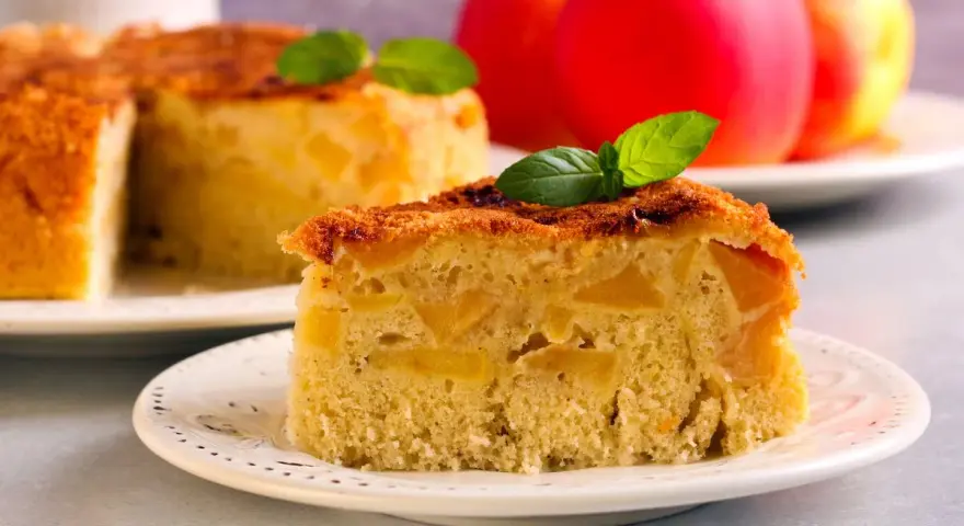 Una fetta di torta di mele vegana, soffice e ricca di mele, guarnita con menta. Sullo sfondo, una torta intera e mele rosse.