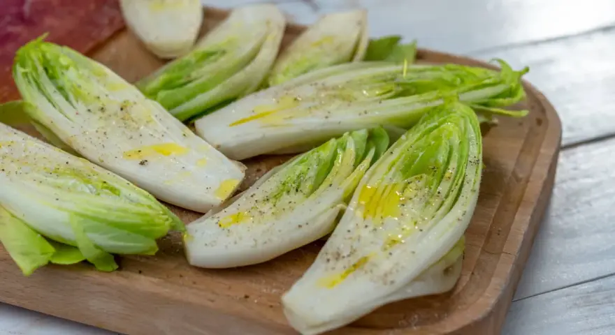 Insalata belga tagliata a metà, condita con olio e pepe, su un tagliere di legno.