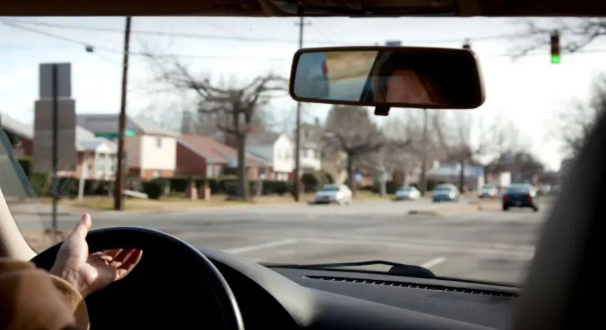 Mano sul volante, pronta per una manovra di svolta. Riflesso del volto nel retrovisore, case e alberi spogli fuori dal finestrino.