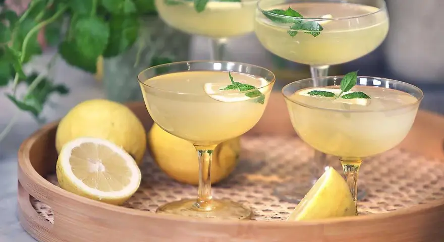 Fyra glas med en ljusgul limoncello drink, garnerade med mynta och citron. Citroner ligger på en träbricka.