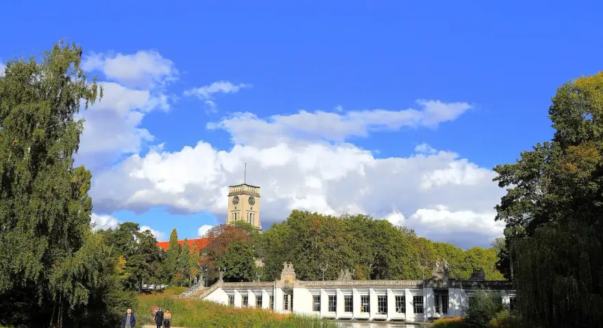 Ein sonniger Tag im **Rudolph Wilde Park**. Ein langer, weißer Pavillon spiegelt sich im Wasser, während ein Uhrenturm über Bäumen aufragt.