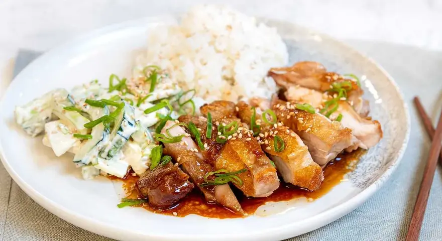 En tallrik med saftig chicken teriyaki, ris och en krämig gurksallad. Garnerad med salladslök och sesamfrön.