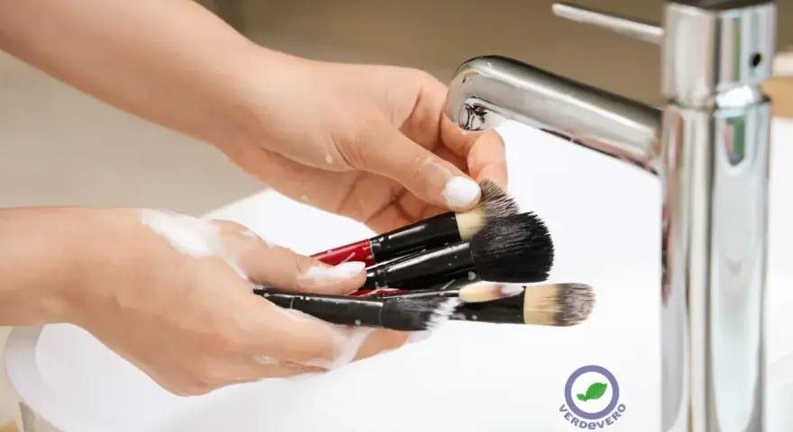 Mani che lavano pennelli da trucco insaponati sotto un rubinetto cromato. Impara come pulire i pennelli da trucco per una pelle sana.