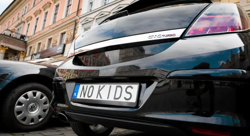 Czarne auto z indywidualnymi tablicami rejestracyjnymi "NO KIDS" na tle kamienicy.