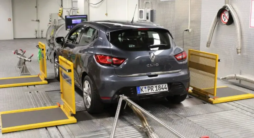 A dark grey Renault Clio is on a dynamometer in a testing facility, with exhaust pipes connected.