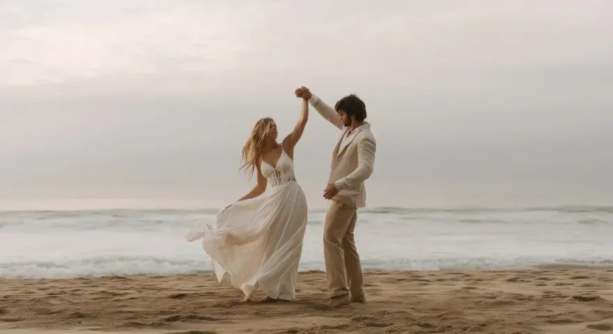 Coppia felice balla sulla spiaggia. Lui indossa un completo chiaro, perfetto per un matrimonio in spiaggia come vestirsi uomo.