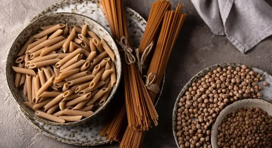 Jasny makaron penne w misce i ciemny makaron spaghetti na talerzu, obok ciecierzycy.