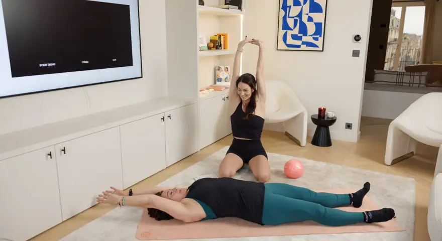 Deux femmes font du sport à la maison, une allongée sur un tapis, l'autre assise. Idéal pour un sport canicule sans sortir.