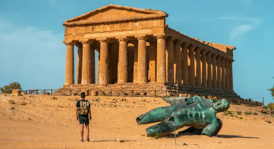 A lone traveler explores the Valley of the Temples in Sicily, standing before an ancient Greek temple and a fallen bronze statue.
