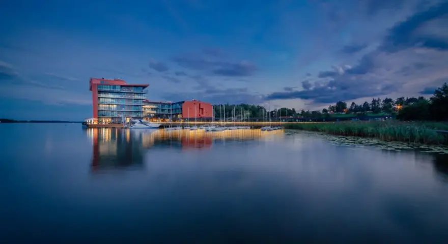 Nowoczesny hotel na ptasiej wyspie, otoczony spokojną wodą i zielenią, z łodziami przy pomoście.