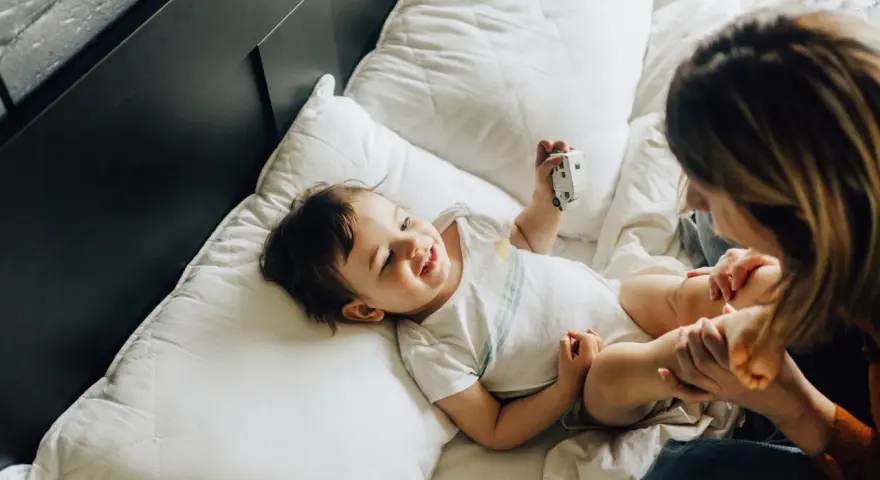 Uśmiechnięte dziecko bawi się zabawką na łóżku. Mama patrzy na malucha. Co to za mastka?