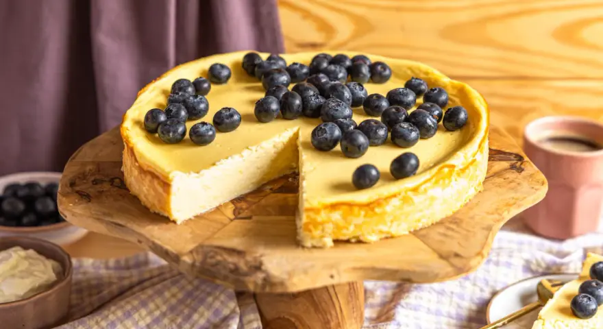 Ein köstlicher Skyr Kuchen, üppig belegt mit frischen Blaubeeren, wartet auf seine Genießer. Daneben eine Tasse Kaffee.