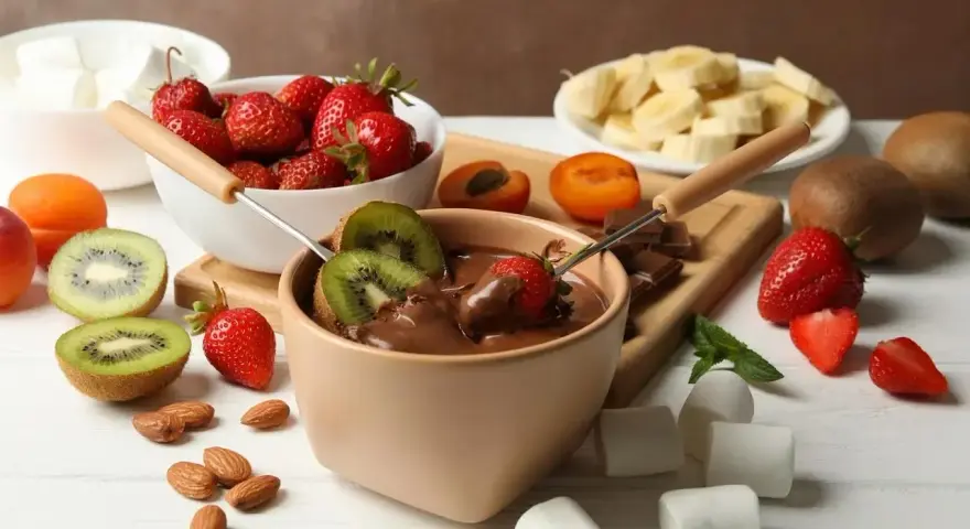 Jak zrobić fondue czekoladowe: zanurzaj truskawki i kiwi w gorącej czekoladzie. Na stole owoce, pianki i migdały.