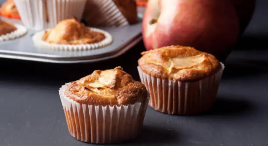 Dos deliciosos muffins de manzana recién horneados, con una manzana roja al fondo.