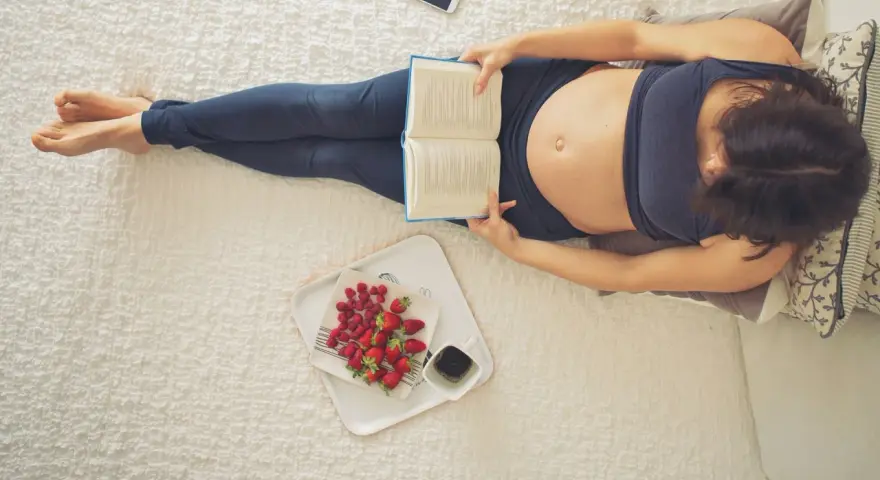 Kobieta w 22 tygodniu ciąży, czyli w 5. miesiącu, relaksuje się z książką i owocami.