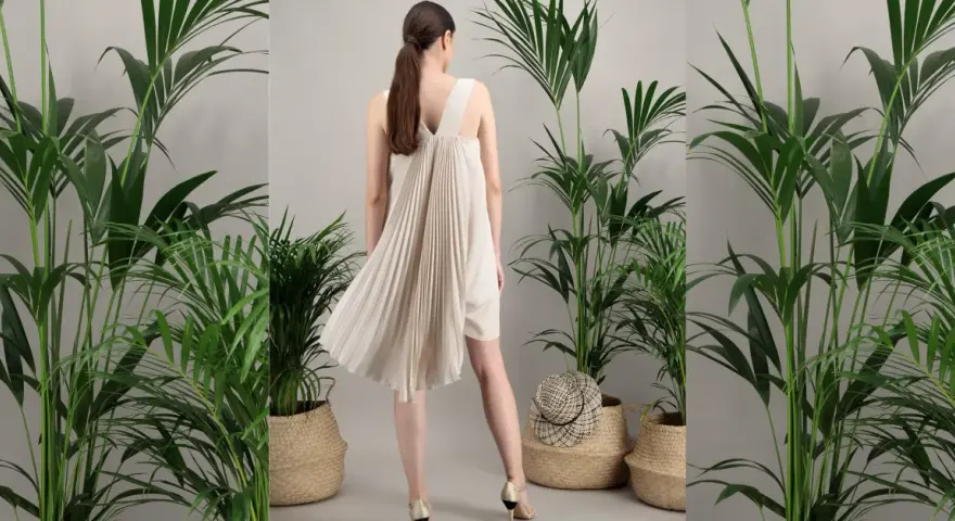 Une femme porte une robe plissée beige, parfaite pour une tenue mariage septembre. Elle est entourée de plantes vertes luxuriantes dans des paniers en osier.