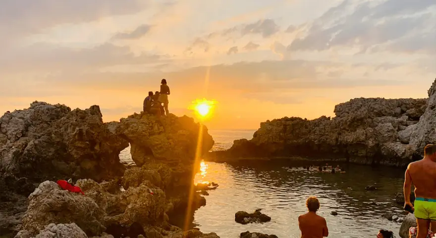 Tramonto dorato sulla piscina di Venere a Milazzo. Persone sedute sulle rocce e in acqua, godendosi la vista.