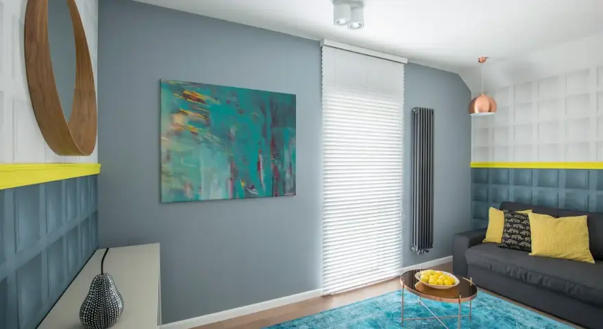 Sala de estar con sofá gris, alfombra turquesa y arte abstracto en tonos fríos. Un espejo redondo de madera y detalles en amarillo complementan el espacio.