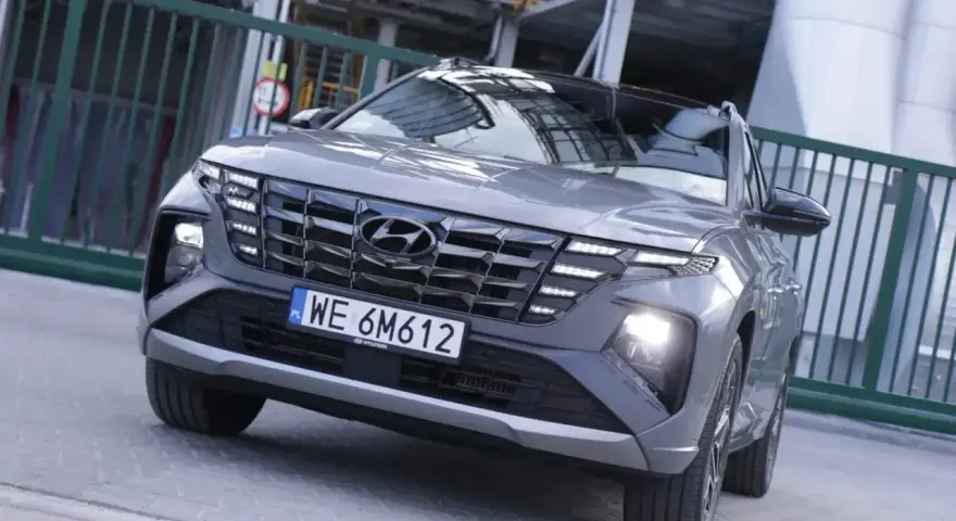 Szary Hyundai Tucson hybryda z charakterystycznym grillem i światłami LED, zaparkowany przed zielonym ogrodzeniem.