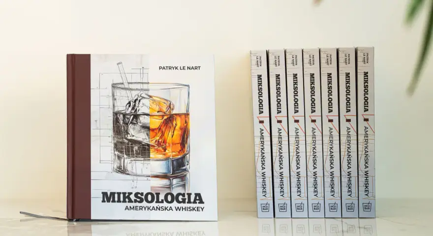 Książka "Miksologia Amerykańska Whiskey" Patryka Le Narta, z ilustracją szklanki z lodem i bursztynowym trunkiem. Obok niej rząd identycznych tomów.
